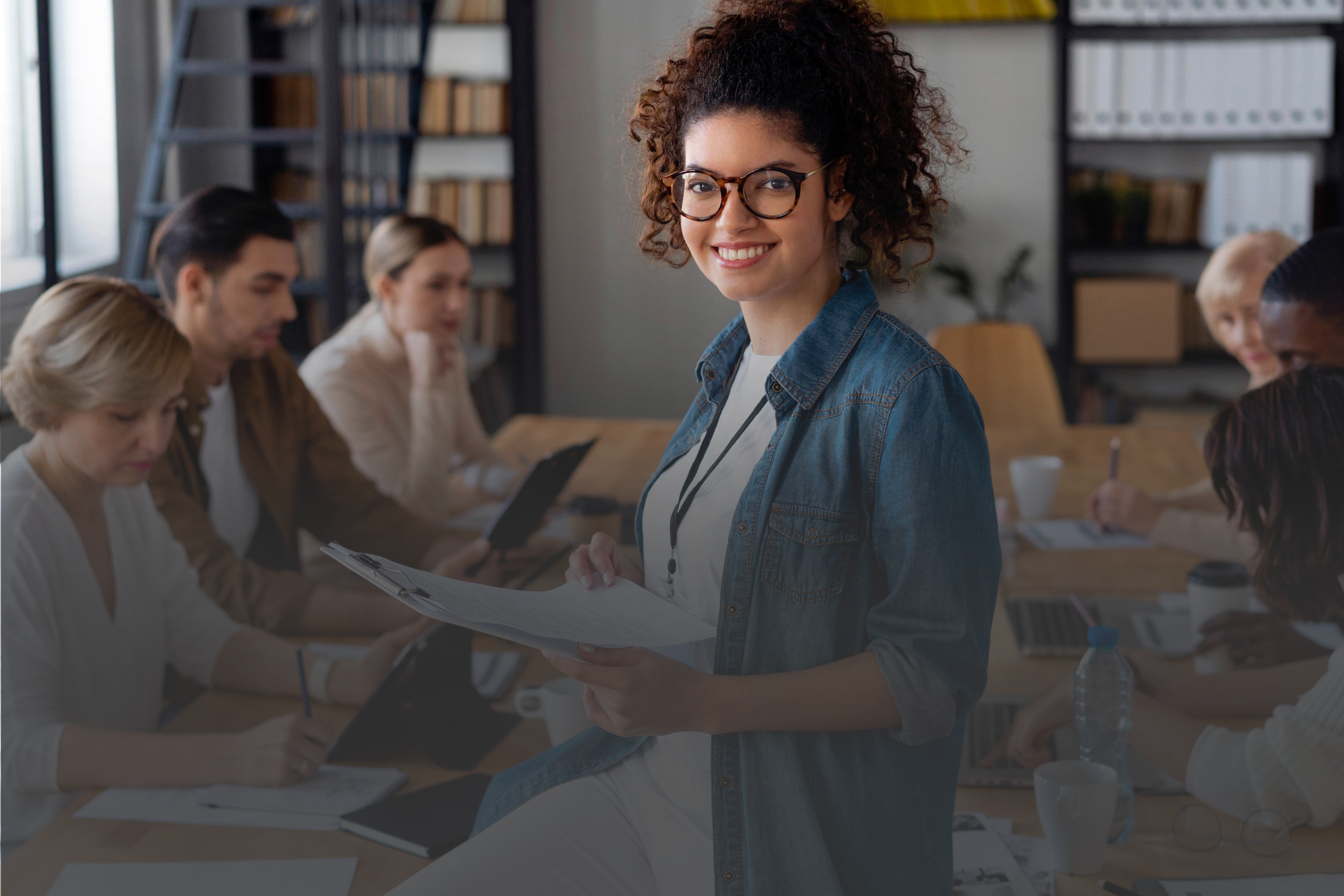 várias pessoas em um ambiente corporativo sentadas em uma mesa de reunião com uma mulher em pé no centro da imagem 