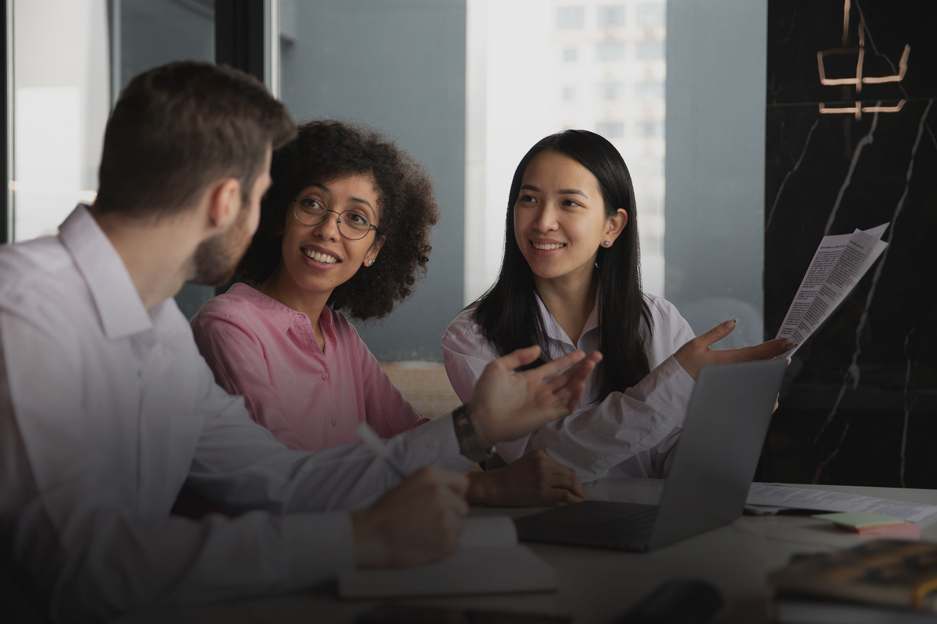 Tres pessoas em um ambiente corporativo conversando sentadas em uma mesa, com papeis e notebook. 