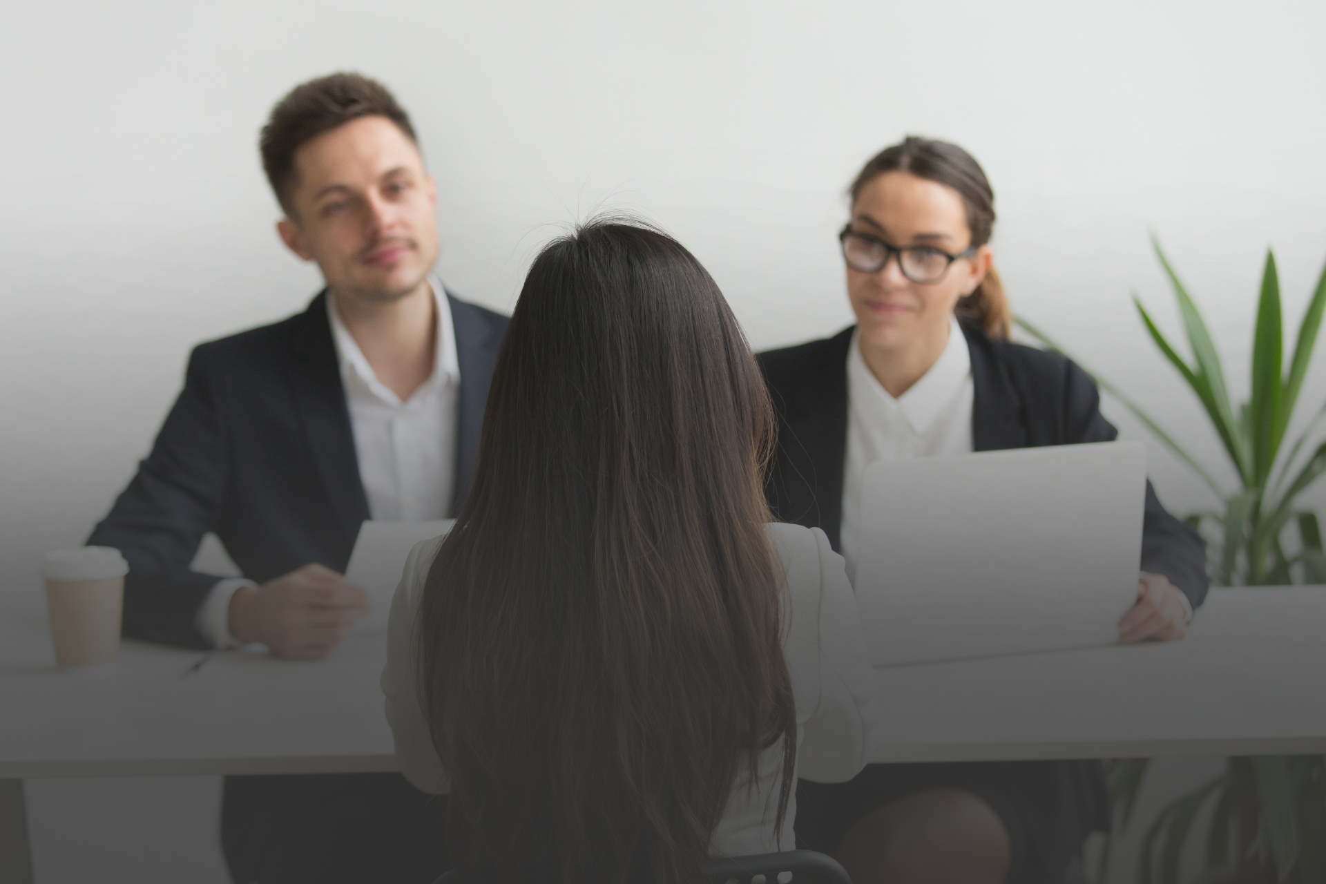 Duas pessoas sentadas de um lado da mesa em um ambiente corporativo olhando para uma pessoa que está do outro lado da mesa, de costas para a foto